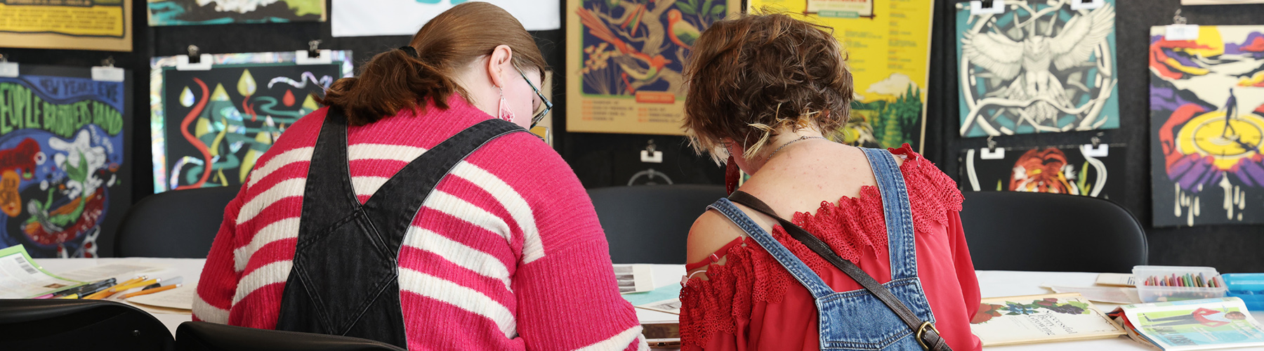 Image of two people seated at a table with art in the background working on a blackout poem at NOTSTOCK